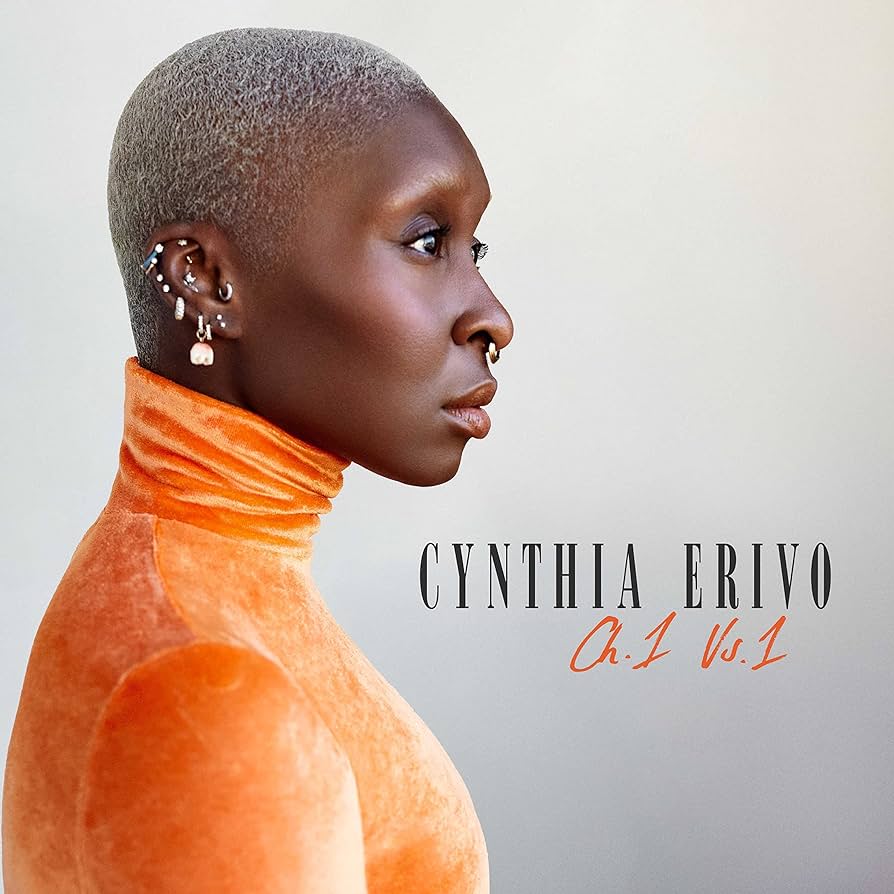 A person with a shaved head wearing an orange top and makeup, with the text 'CYNTHIA ERIVO CH. 1 Vs.